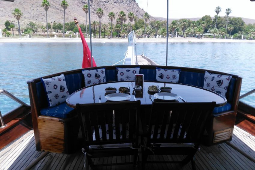 Bodrum Bitez tekne kiralama turlarında misafirler için özenle hazırlanan taze Ege mezeleri ve zeytinyağlılarla donatılmış; masmavi deniz manzarasına karşı ahşap arka güverte yemek masası.