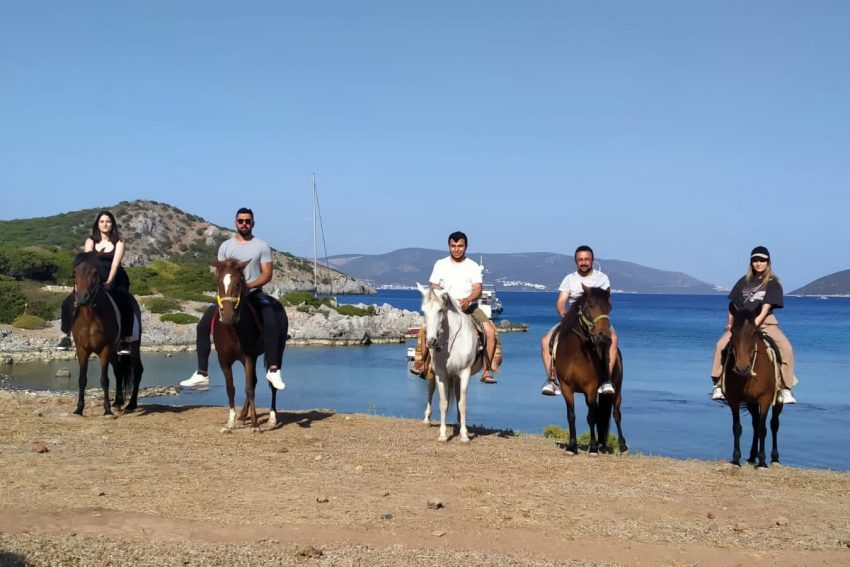 Koy ve tekne manzaraları eşliğinde doğayla iç içe Bodrum at safari turu; sahil kenarında açık havada at binme keyfi yaşayan mutlu turistler.