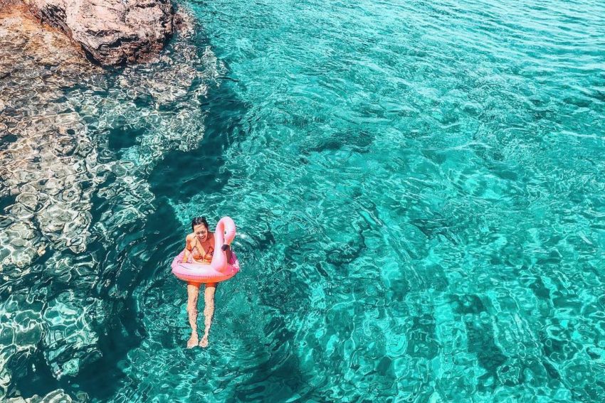 Gümbet Tekne Tur Gezisi.Kayalık bir koyda, cam gibi berrak turkuaz suların içinde pembe flamingo deniz simidiyle yüzen ve gülümseyen kadın.
