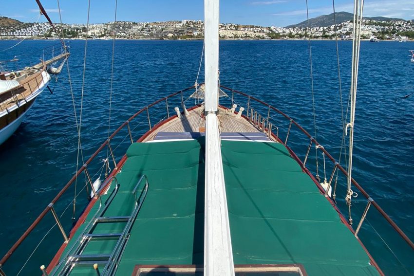 Bodrum tekne kiralama turları için ideal, geniş yeşil güneşlenme minderlerine sahip geleneksel ahşap guletin ön güvertesinden panoramik Bodrum sahili ve masmavi deniz manzarası.