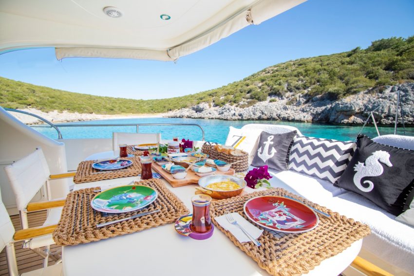 Bodrum Torbada Turkuaz sulara sahip ıssız bir koyda, lüks yatın arka güvertesinde hazırlanan zengin Serpme Türk Kahvaltısı ve deniz manzarası.